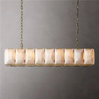 Linear chandelier with brass finish and stacked alabaster stone panels suspended by chains against a gray wall.